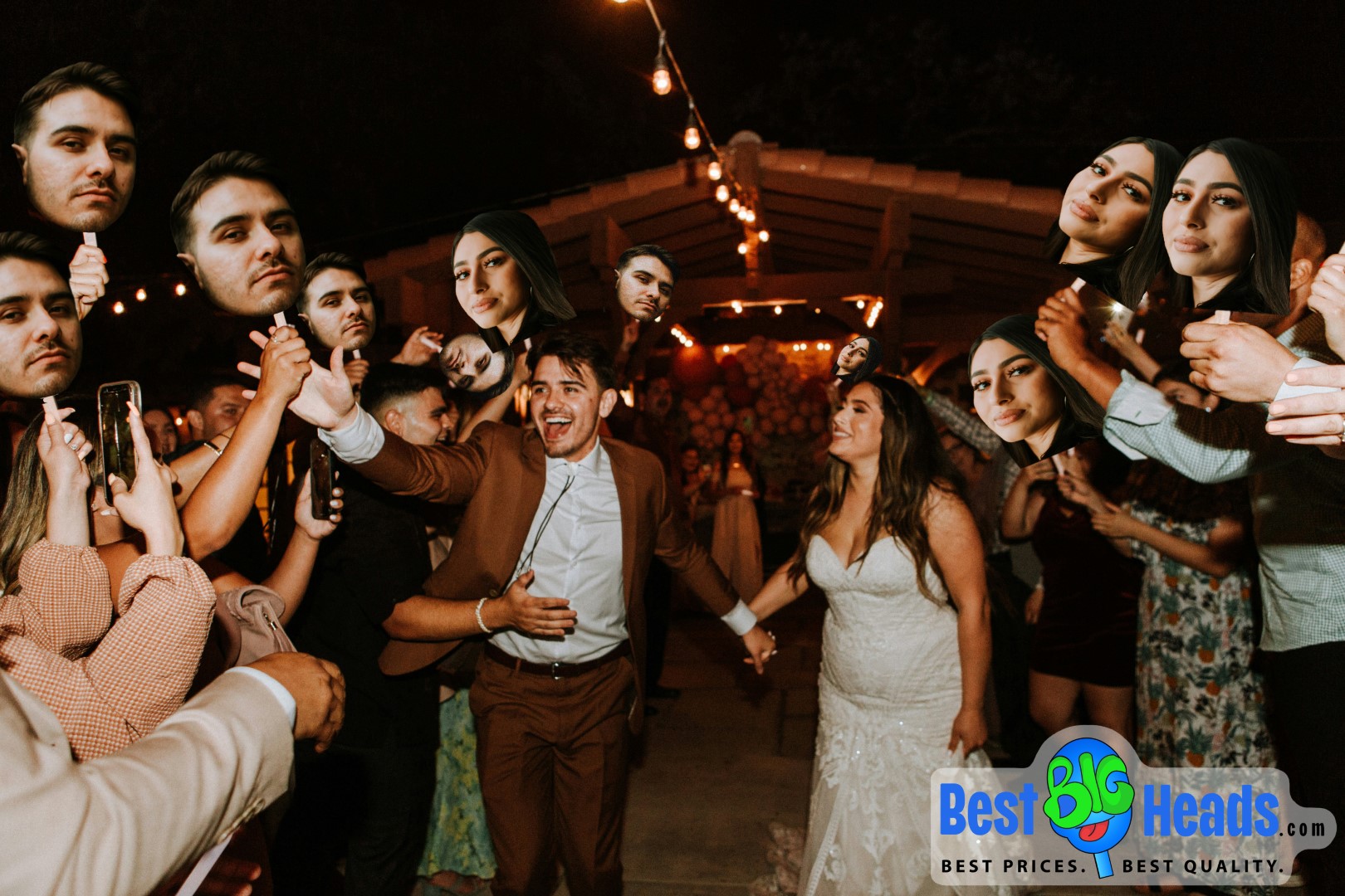 A newlywed couple is joyfully walking through a crowd of guests at their outdoor wedding reception. The nighttime setting is beautifully lit with string lights, creating a warm and celebratory atmosphere. The guests are enthusiastically holding up big head cutouts of both the bride and groom, adding a fun and personalized touch to the celebration. The couple is all smiles, holding hands, and clearly enjoying the moment. The "Best BIG Heads™" logo is visible at the bottom right corner, showcasing the source of the custom big head cutouts.